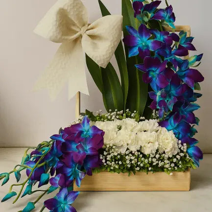 Blue orchids and white flowers arranged in a white cylindrical container with a bow on top.