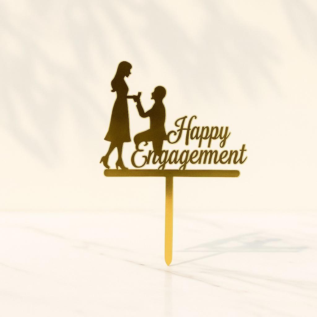 Gold 'Happy Engagement' cake topper with a silhouette of a man proposing to a woman on a white background.